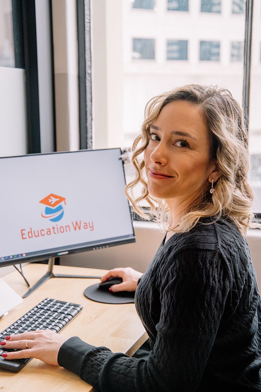 Mulher sentada à mesa, olhando para a câmera. O monitor do computador exibe o logotipo "Education Way".