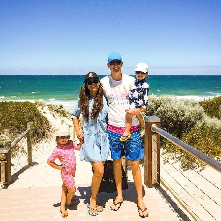 Família de quatro pessoas em um calçadão na praia; dia ensolarado. Mulher de vestido azul, homem segurando criança, criança pequena de maiô.
