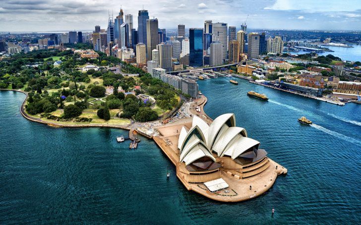 A Ópera de Sydney, na Baía de Sydney, Austrália, com o horizonte da cidade ao fundo e barcos na água.