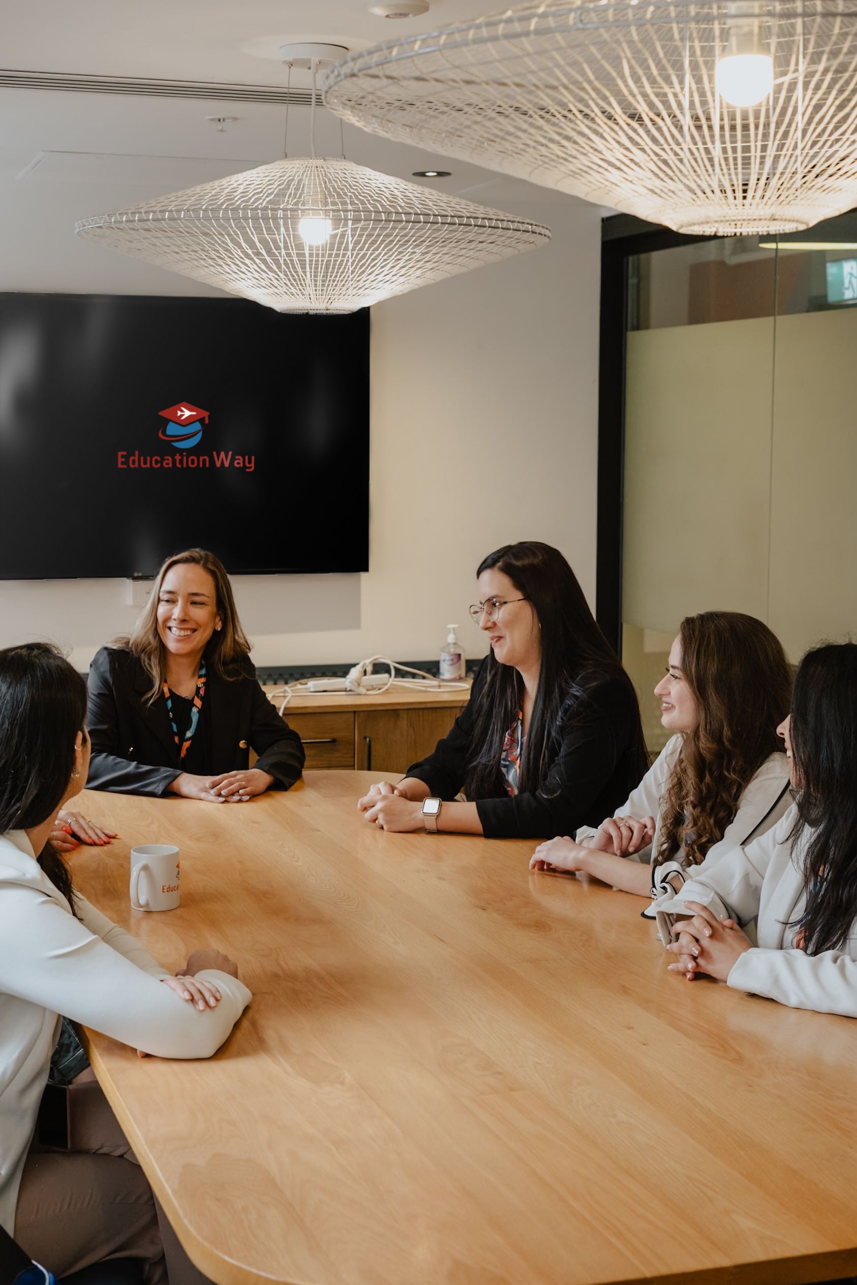 Pessoas em volta de uma mesa de conferência de madeira em um escritório, discutindo enquanto um logotipo é exibido na tela atrás delas.