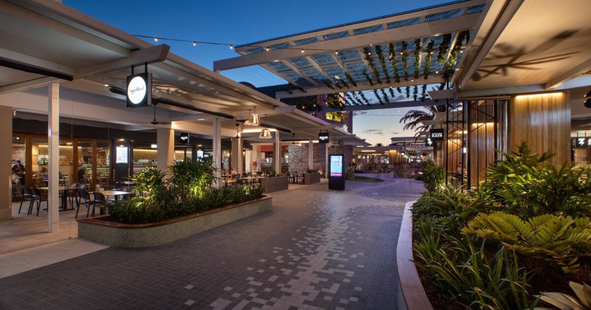 Passarela ao ar livre de um shopping center com restaurantes e plantas decorativas, à noite.
