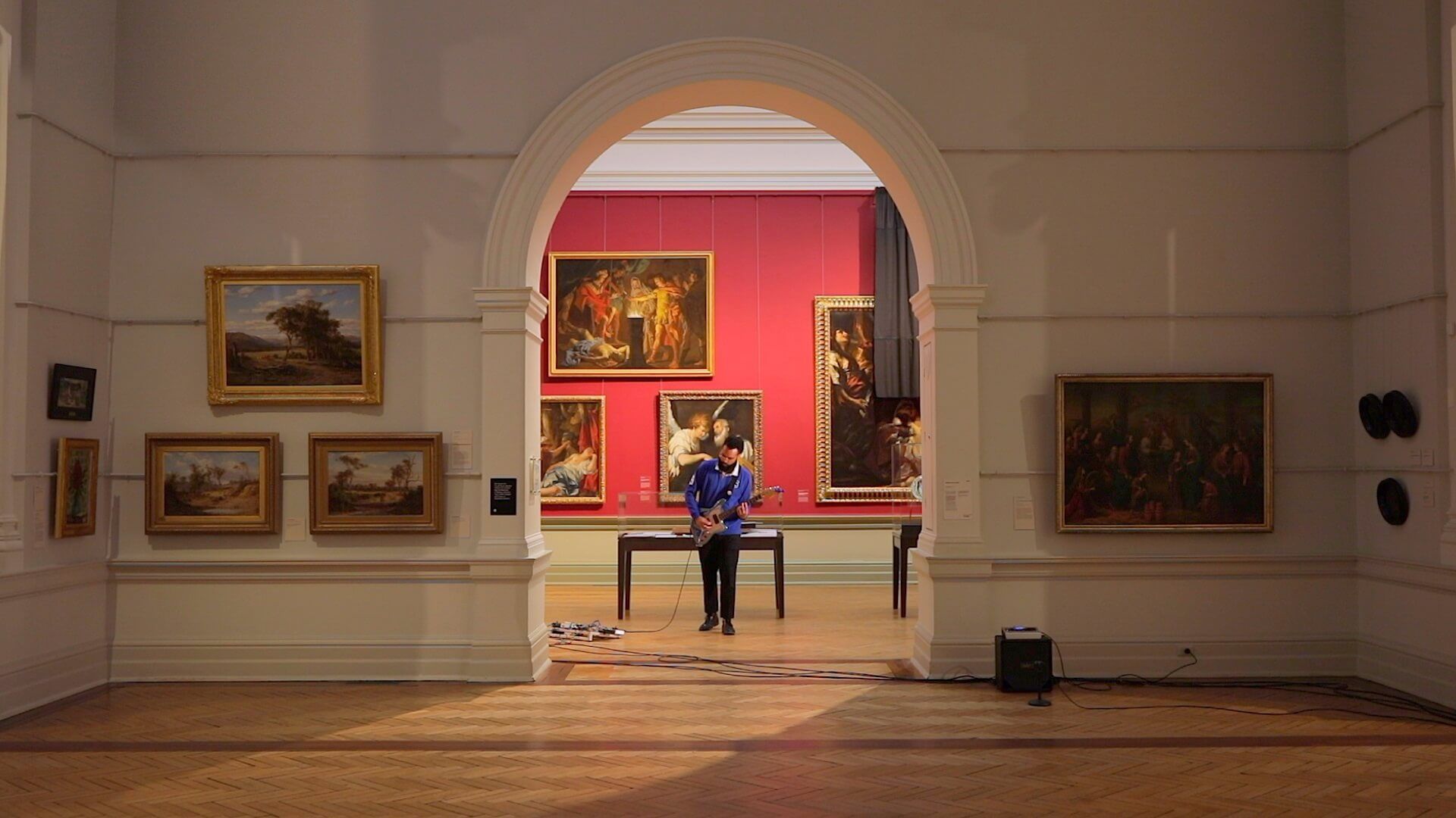 Interior do museu com porta em arco emoldurando pinturas em paredes vermelhas; pessoa de jaqueta azul em pé.