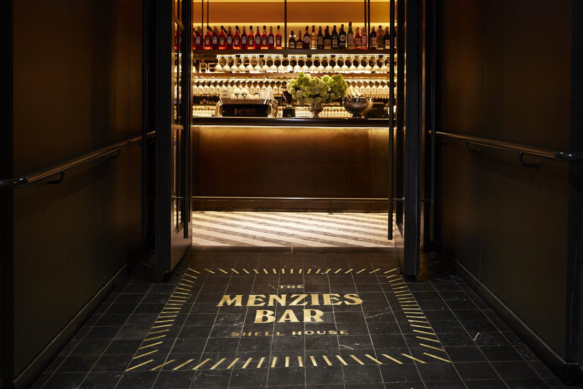Entrada escura para o The Menzies Bar com o bar iluminado ao fundo e texto dourado no piso de azulejos.