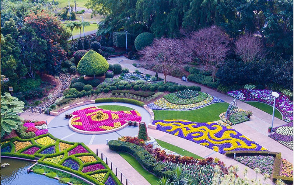 Vista aérea de um jardim colorido com canteiros de flores com padrões e sebes bem cuidadas, situado num ambiente semelhante a um parque.