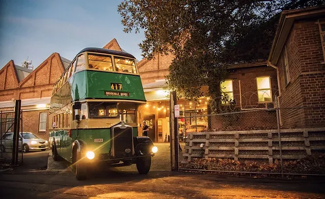 Um ônibus antigo de dois andares, na cor verde, sai por um portão e está estacionado ao lado de um prédio de tijolos ao entardecer.