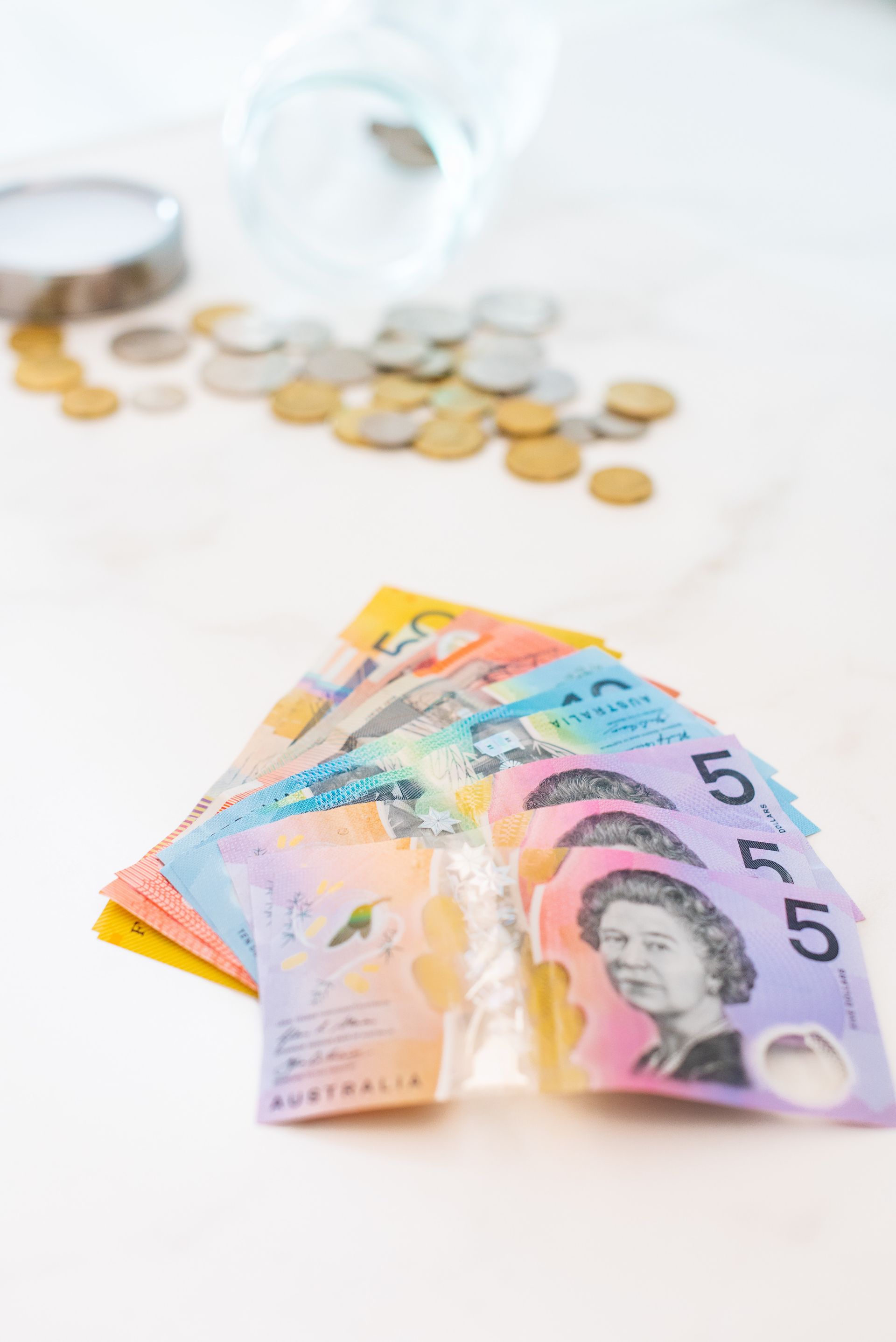 Notas e moedas australianas, derramadas de um frasco, sobre uma superfície branca.