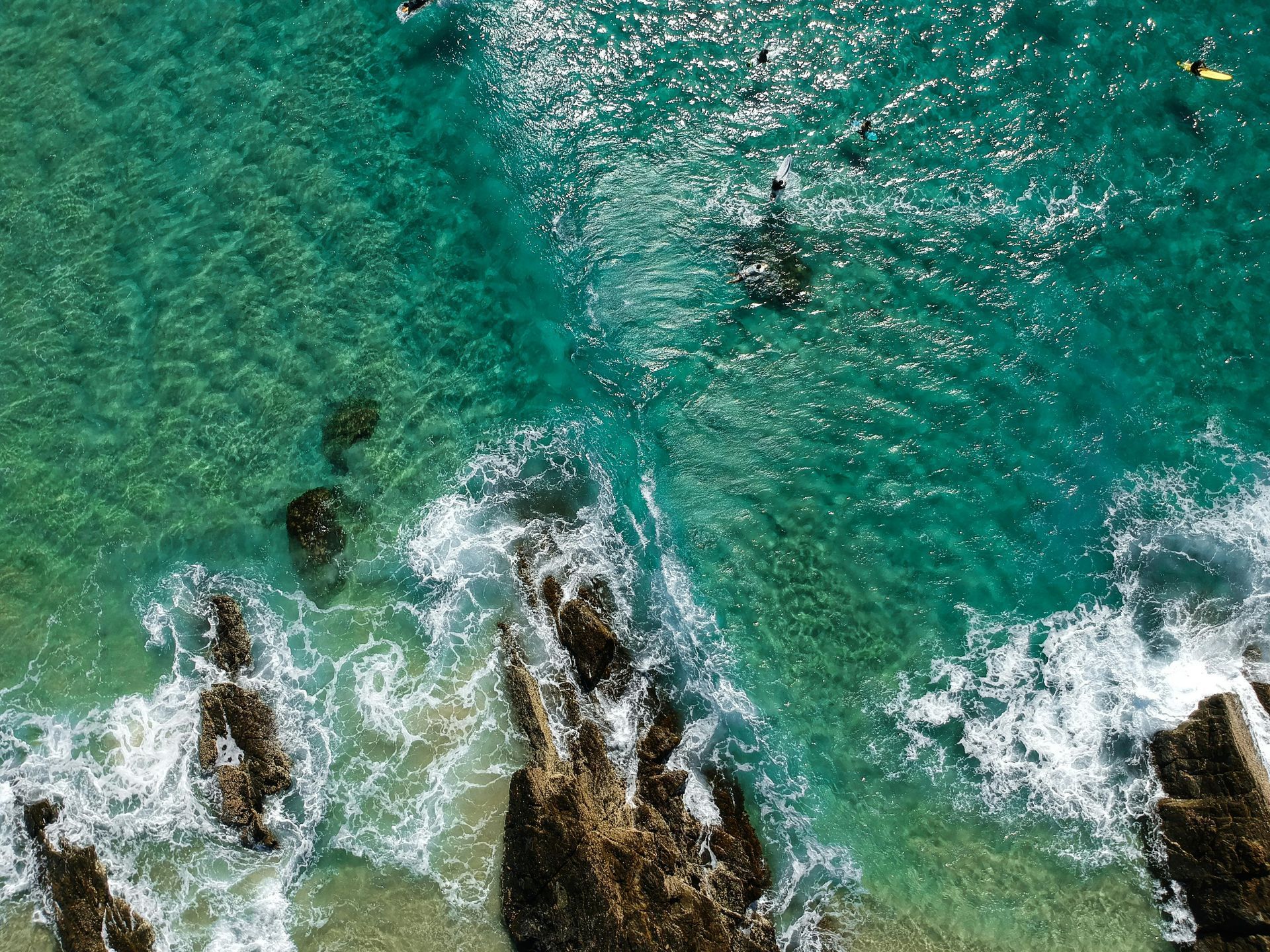 Vista aérea de águas cristalinas azul-turquesa com ondas quebrando em rochas escuras e alguns nadadores.