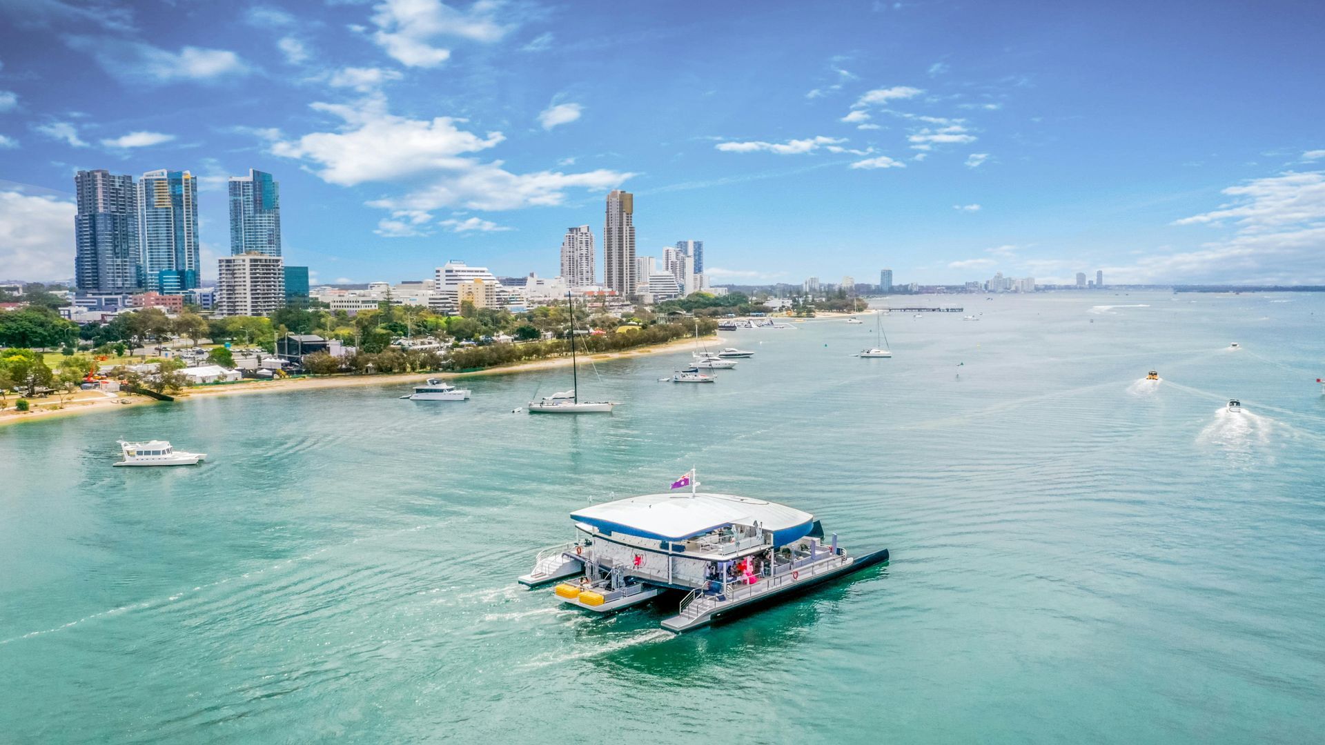 Um catamarã navega em águas turquesas perto da orla de uma cidade com barcos e prédios altos.