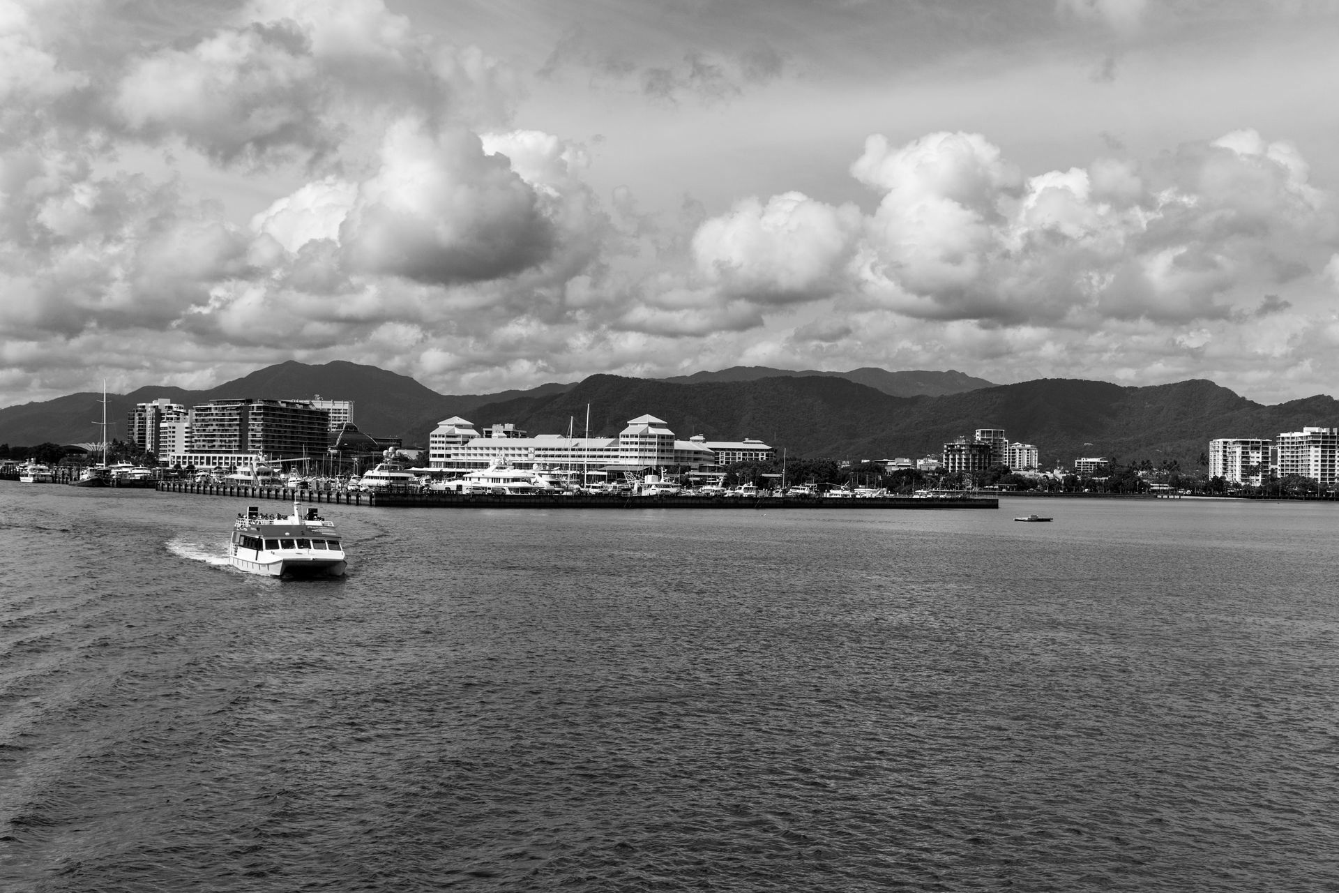 Imagem em preto e branco de uma massa de água com um barco à vela, edifícios da cidade e montanhas ao fundo.