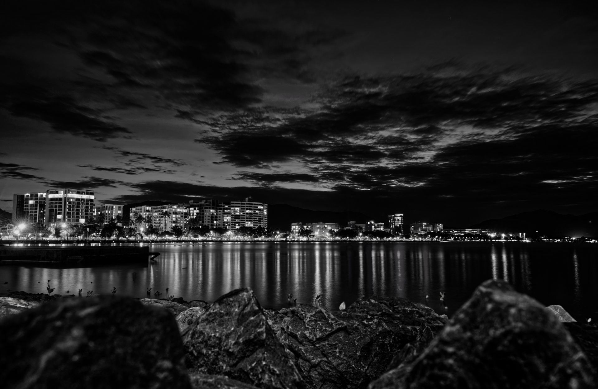 Paisagem urbana em preto e branco à noite, com luzes refletidas na água sob um céu nublado. Primeiro plano rochoso.