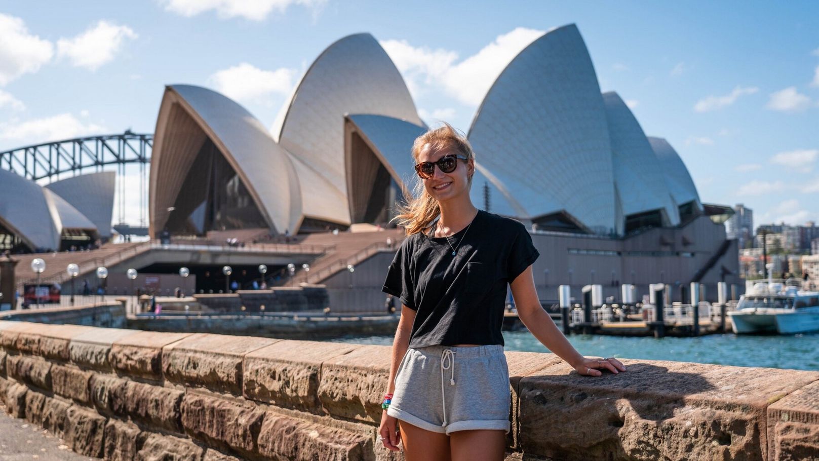 Mulher posando em frente à Ópera de Sydney, usando óculos de sol, blusa preta e shorts cinza.
