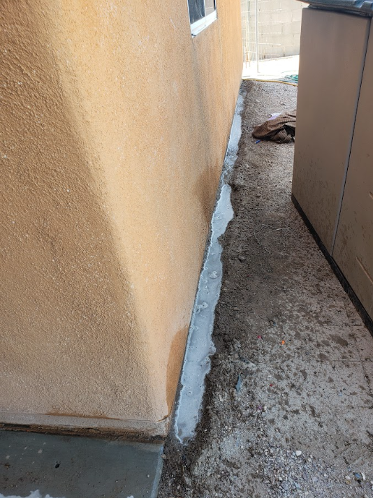 Narrow side yard beside a stucco wall and shed, with a concrete strip and gravel ground.