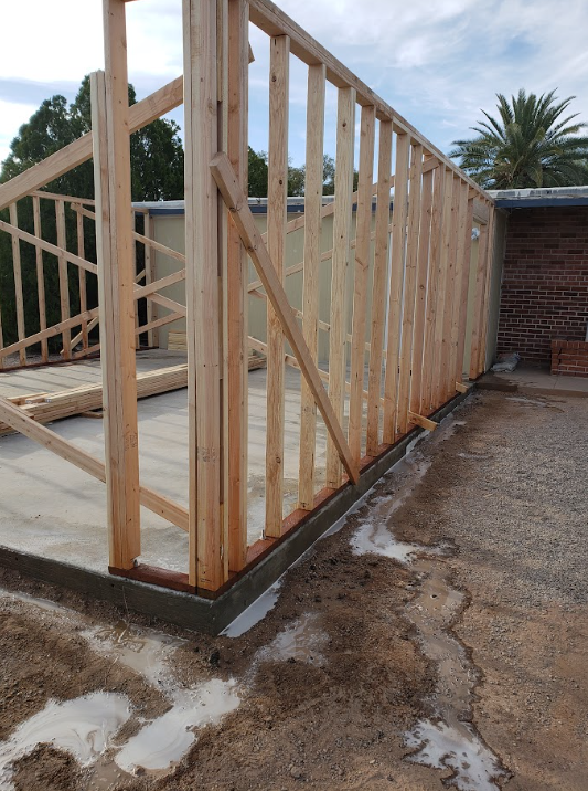 Wooden house frame under construction on a concrete slab, with diagonal bracing and a brick wall nearby