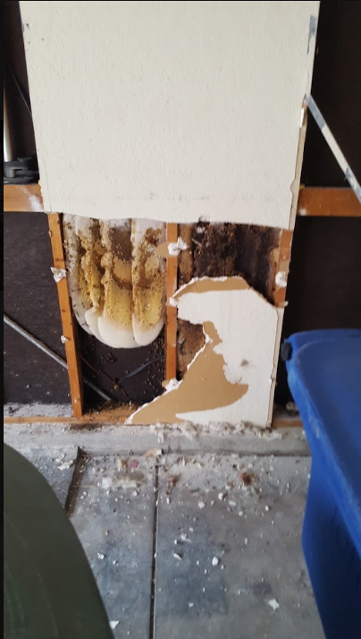 Damaged white wall with exposed insulation and broken paneling near a floor, in a cluttered indoor space