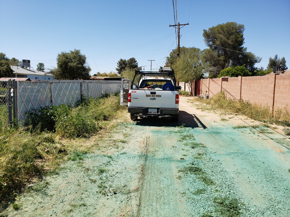 White pickup truck parked on a narrow dirt driveway beside a chain-link fence and brick wall in bright sunlight