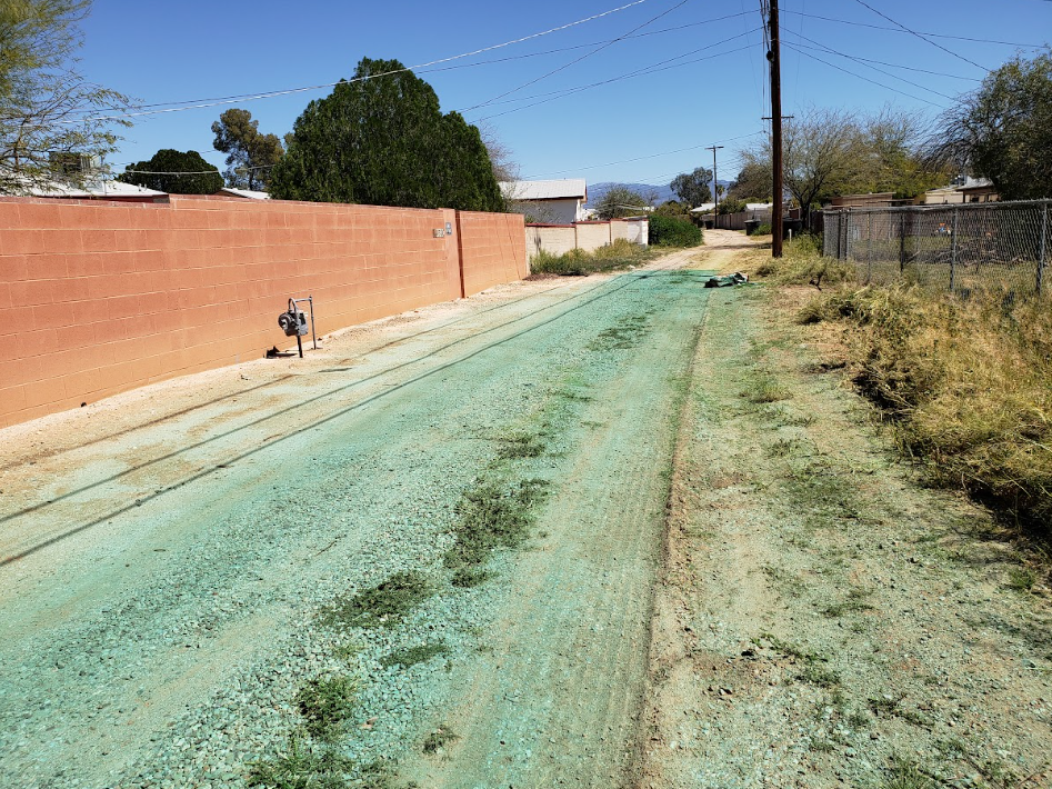 Narrow dirt road with green dust, brick wall on left, chain-link fence and dry grass on right under clear sky