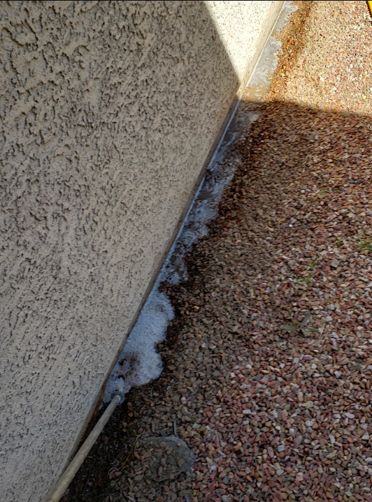 Close-up of a stucco wall beside gravel, with white foam or sealant along the base