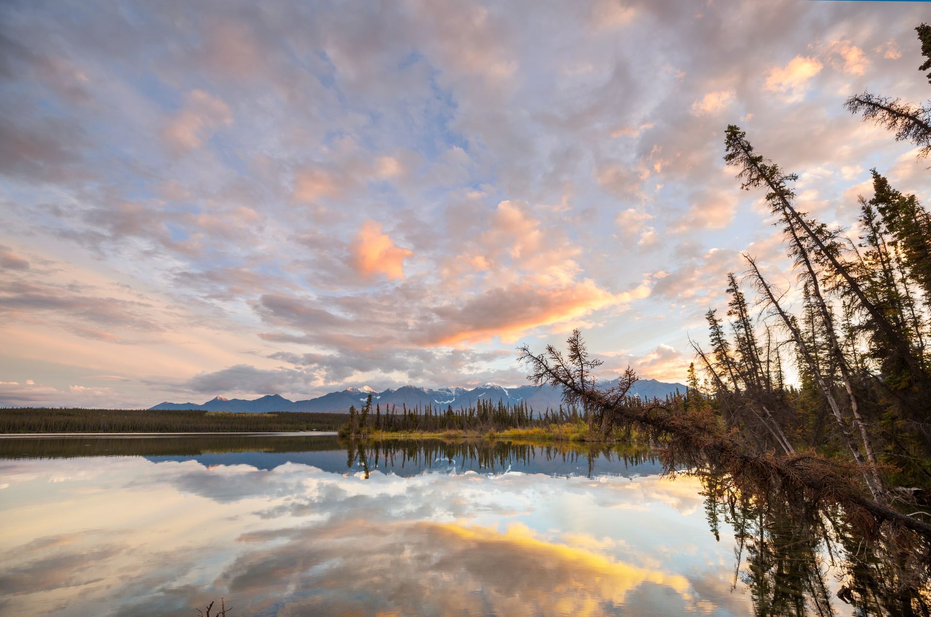 Calm lake reflecting a colorful sunset with clouds, trees, and distant mountains.