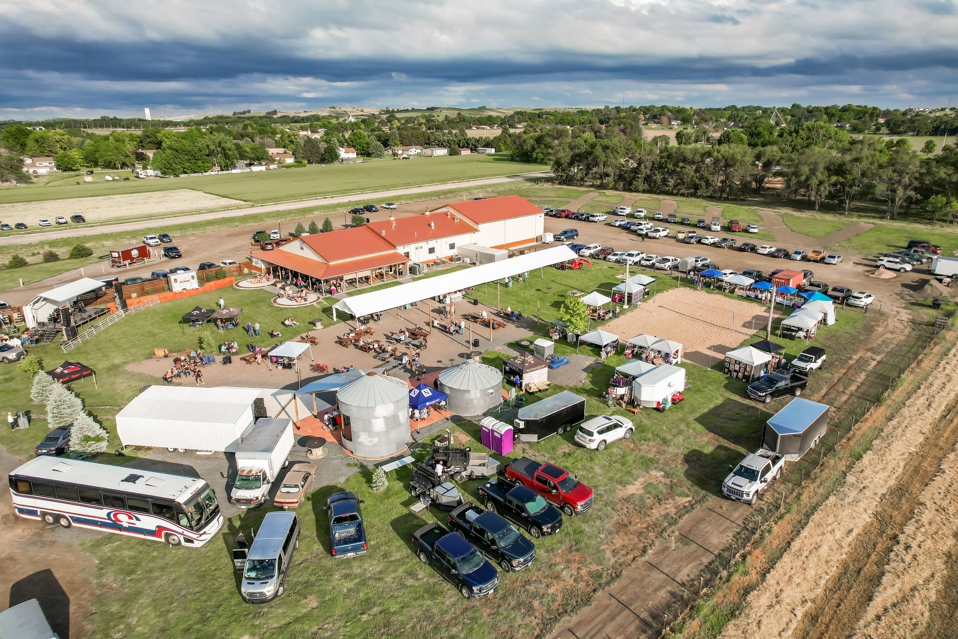 Aerial view of an outdoor event. Building, tents, parked vehicles, and people gathered in a grassy field under a cloudy sky.