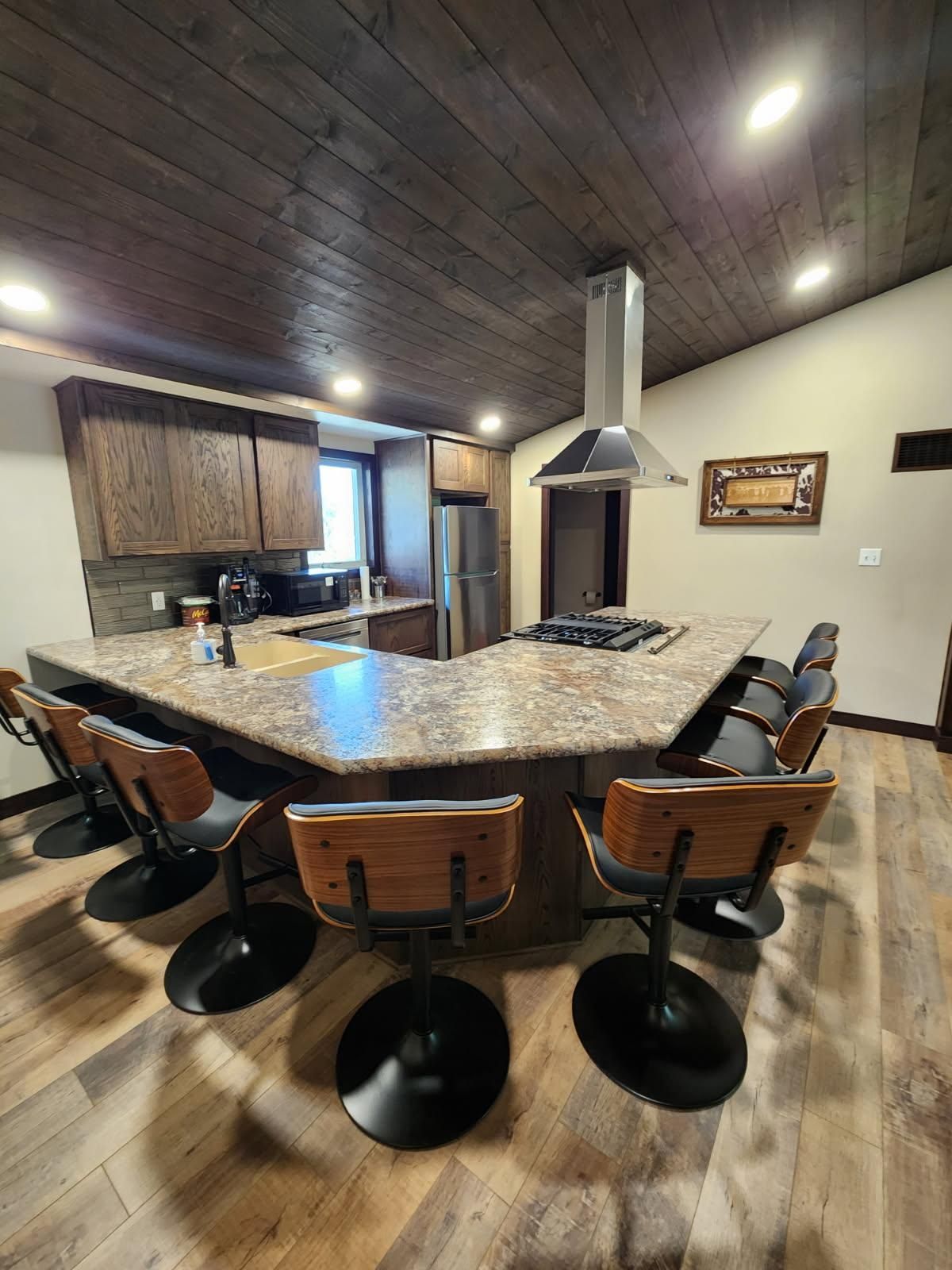 Kitchen with island seating, stainless steel range hood, wood ceiling and cabinets, and light wood flooring.