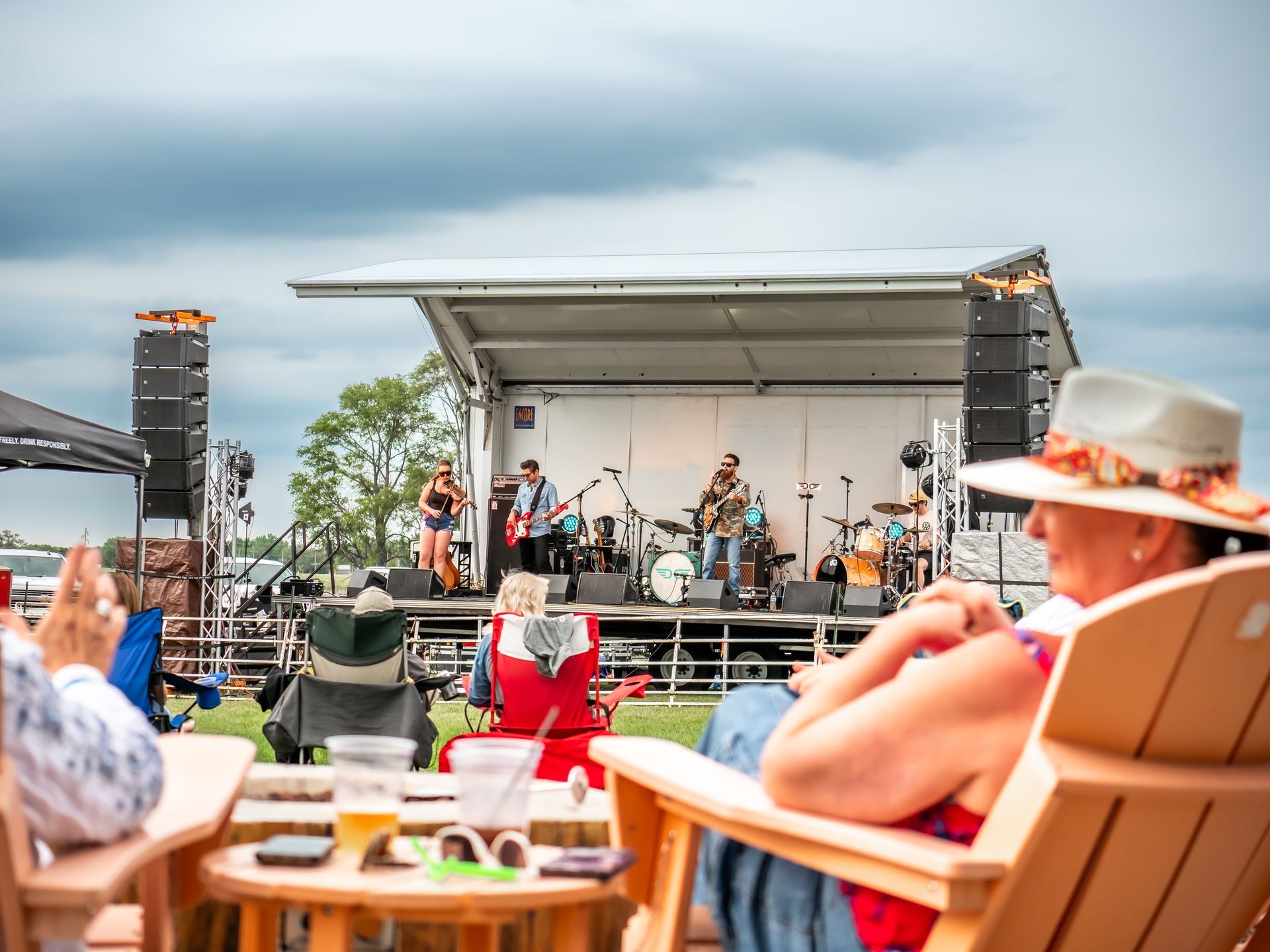 Band performs on outdoor stage, audience seated in chairs, cloudy sky.