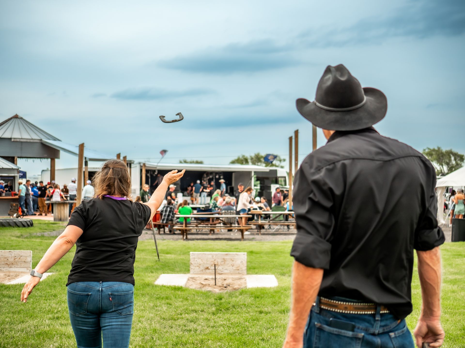 Woman throws horseshoe, man in cowboy hat watches outdoors. People and food stand in background.