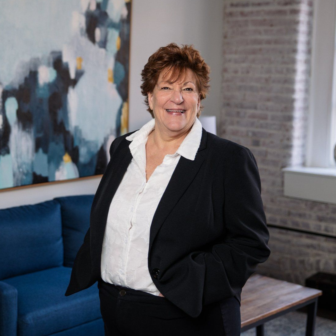 Smiling person in a black blazer stands in a modern office with a blue couch and abstract wall art