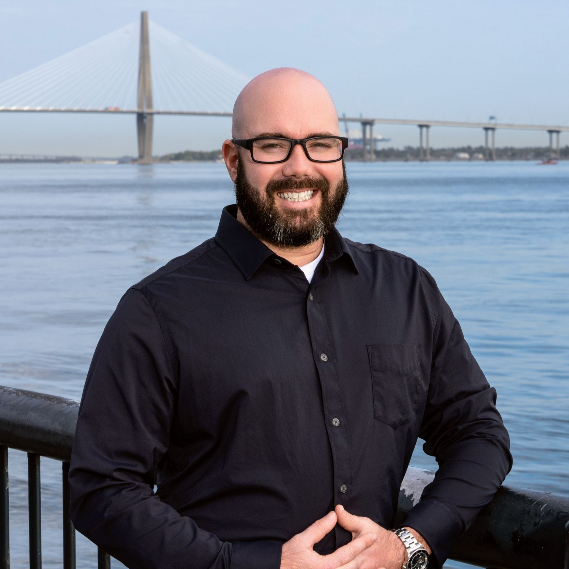 Smiling bald man in black shirt by a waterfront railing with a bridge in the background