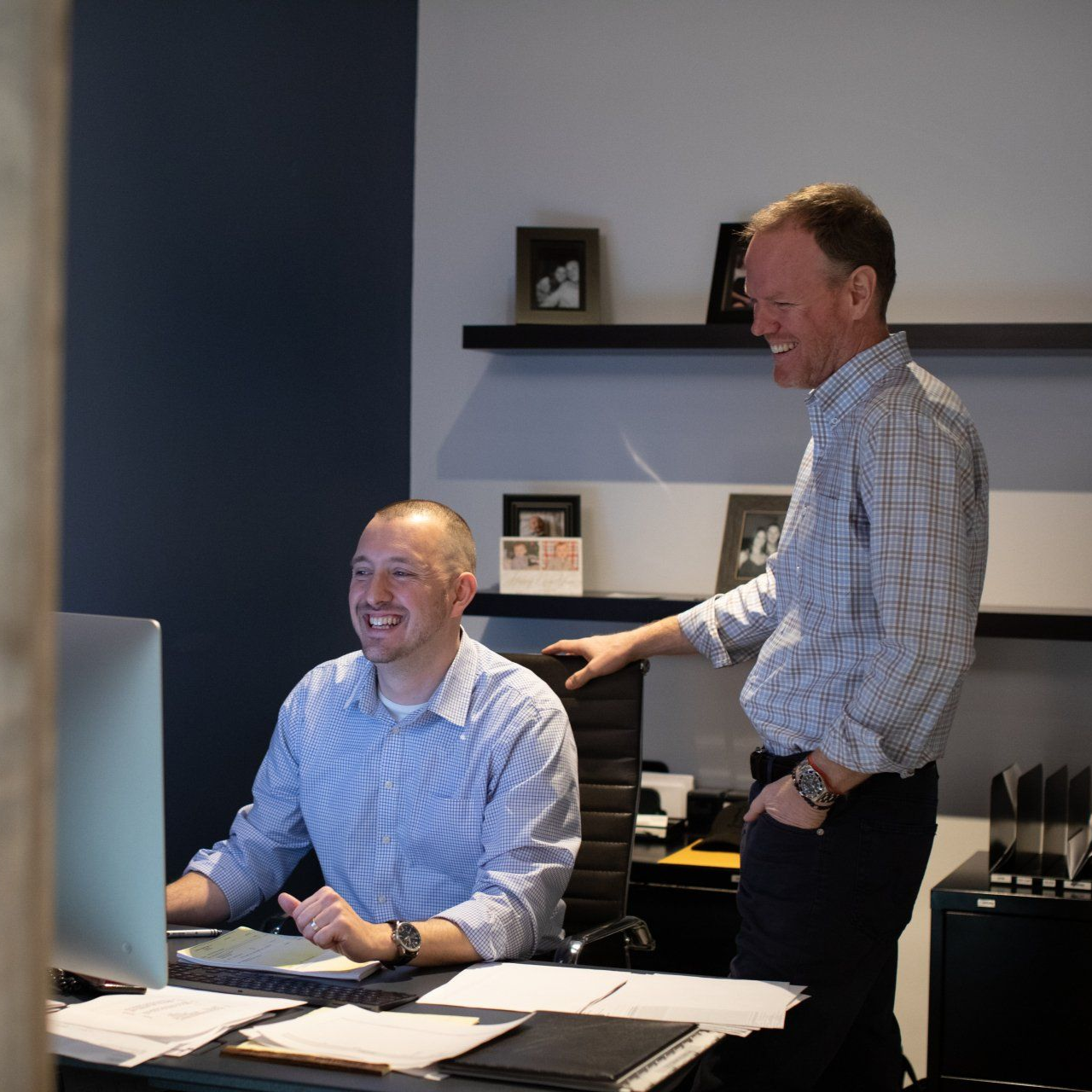 Two coworkers smiling in an office, one seated at a desk and the other standing beside him.
