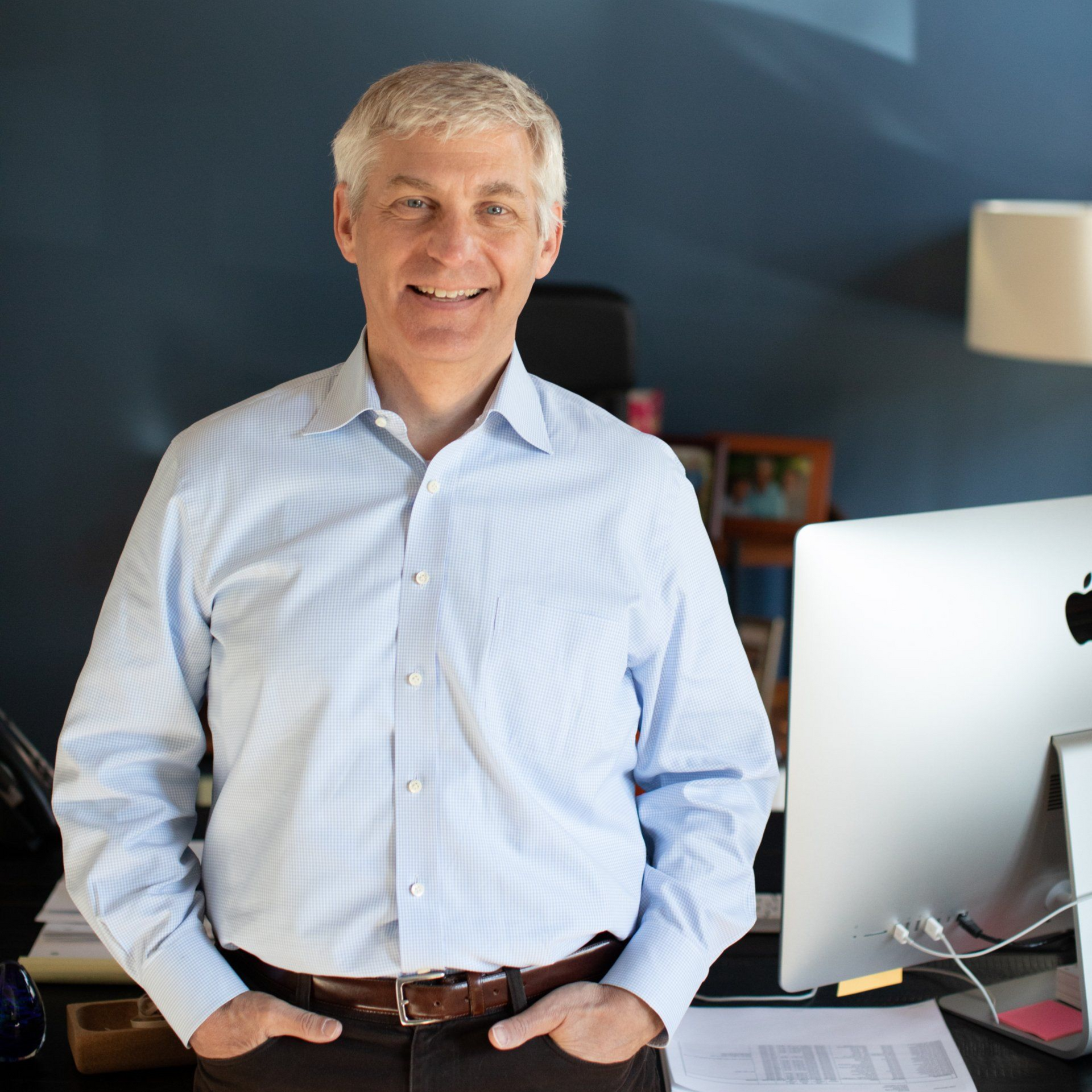 Smiling man in a light blue shirt standing in a home office beside an iMac