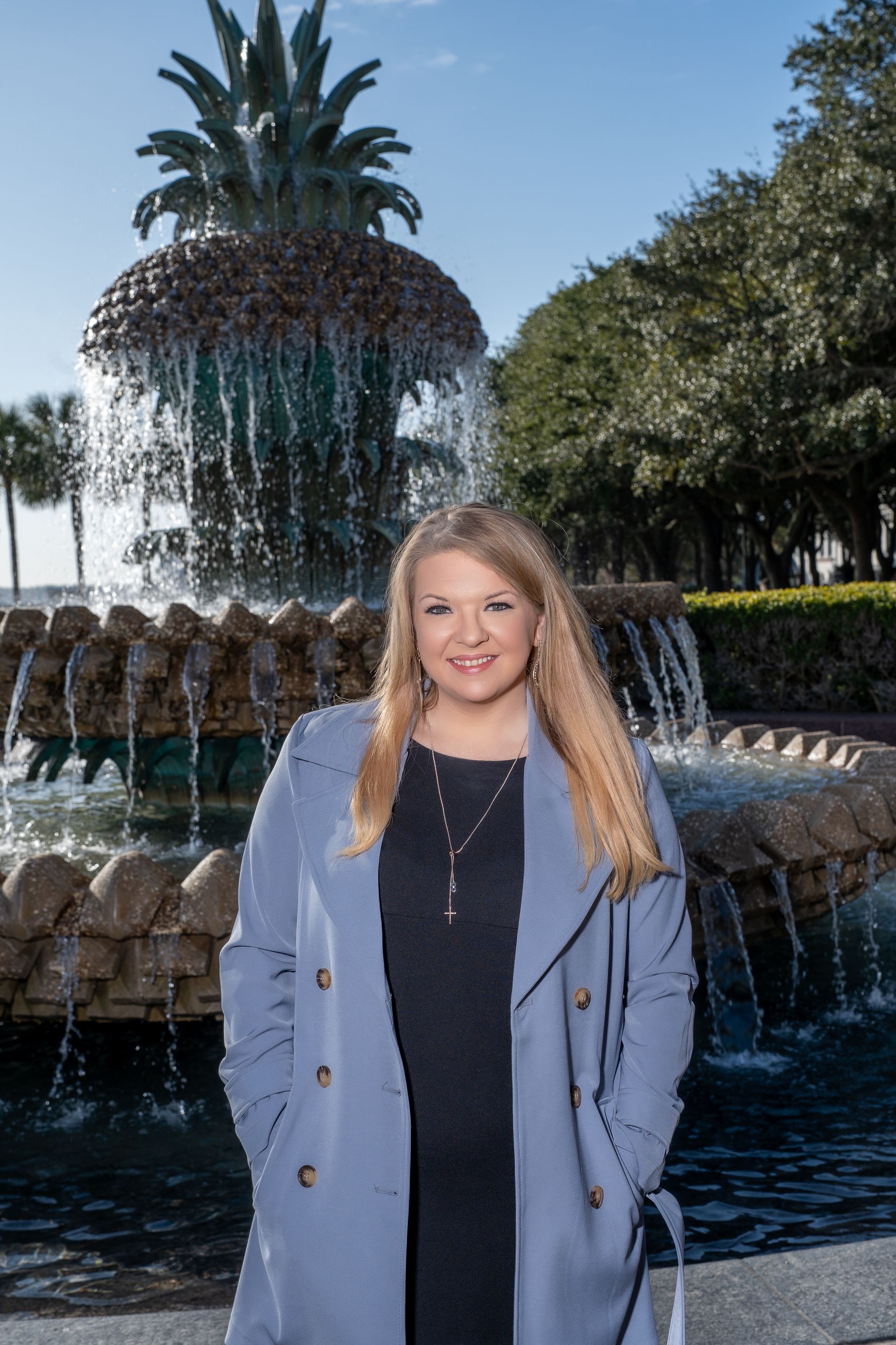 Woman in a blue coat standing in front of a fountain, smiling outdoors in daylight.