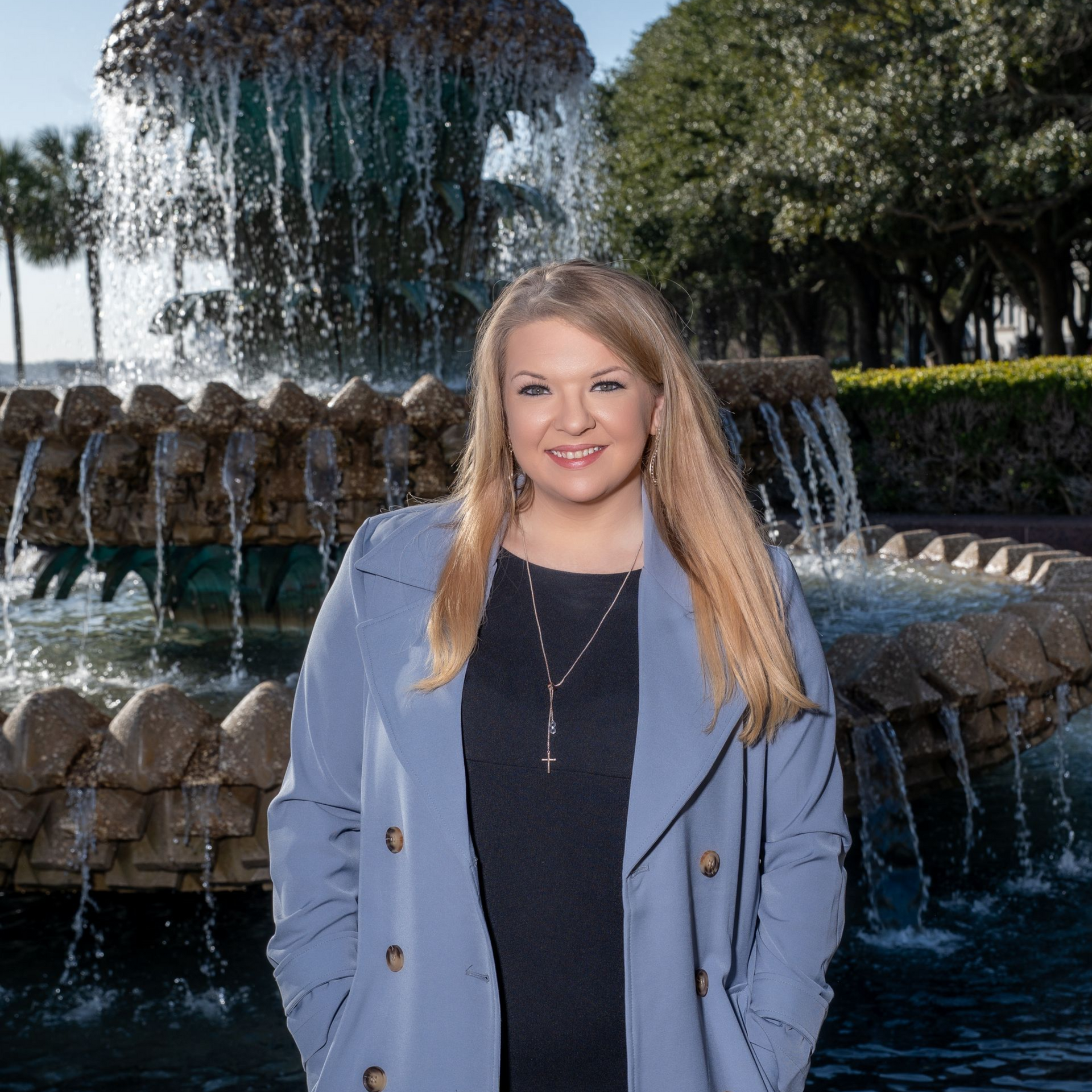 Blonde woman in a blue coat smiling in front of a cascading stone fountain outdoors