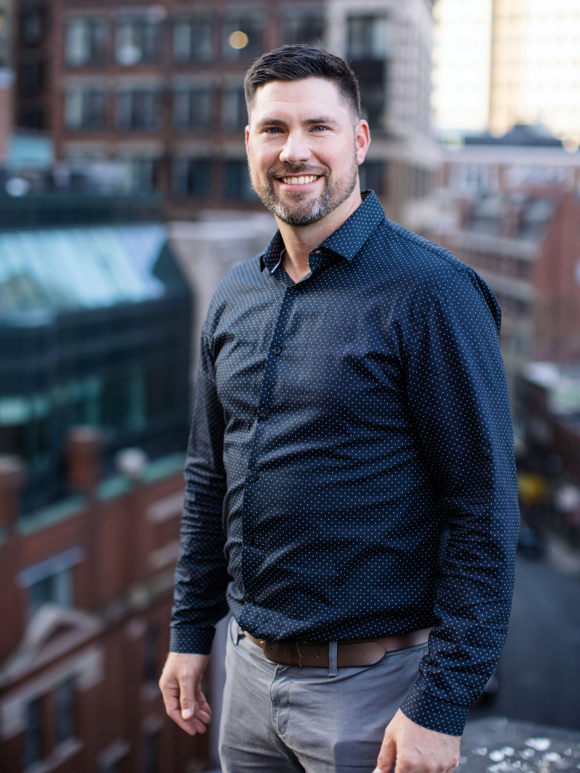 Man standing on a rooftop with city buildings behind him, wearing a navy shirt and gray pants.