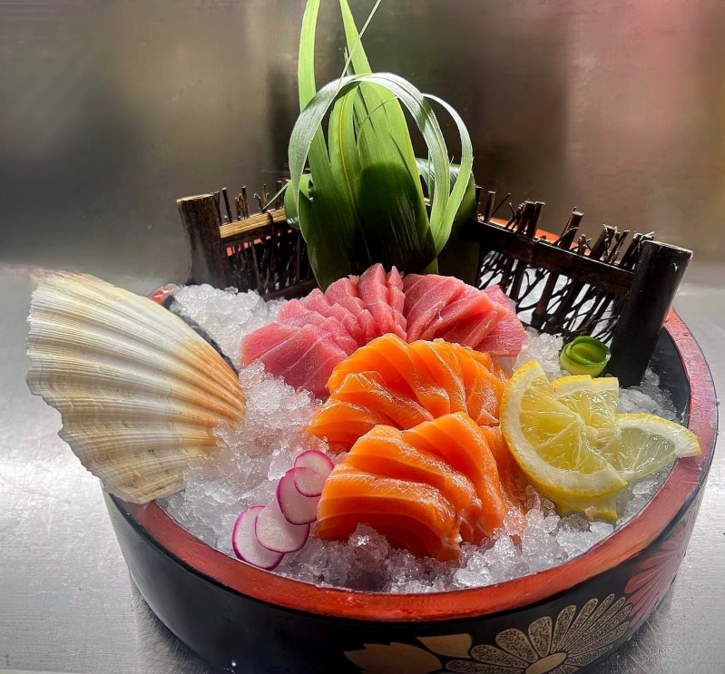 Sashimi assortito e guarnizioni in una ciotola nera su ghiaccio, con capesante, limone e foglie verdi.