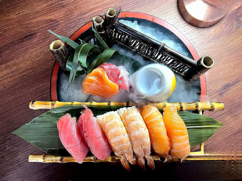 Piatto di sushi assortito su un tavolo di legno, con nigiri su una foglia di banana e foglie decorative sullo sfondo.