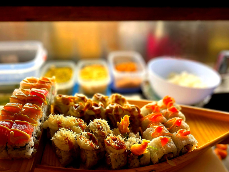 Assortimento di rotoli di sushi su un vassoio di legno con salse e contorni sullo sfondo.