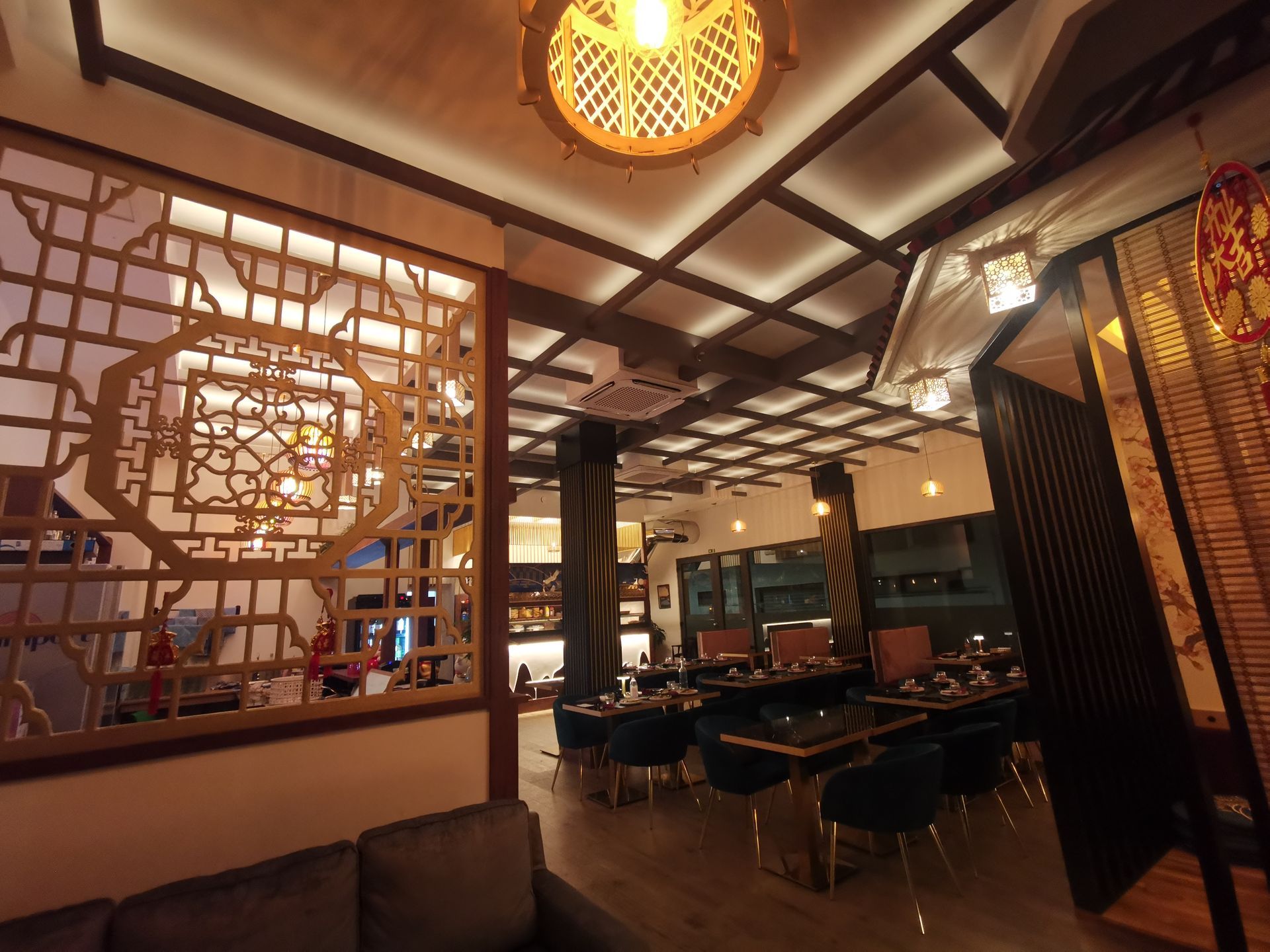 Interno di ristorante illuminato da luci soffuse, con decorazioni in legno a griglia, tavoli e sedie sotto un soffitto decorato.
