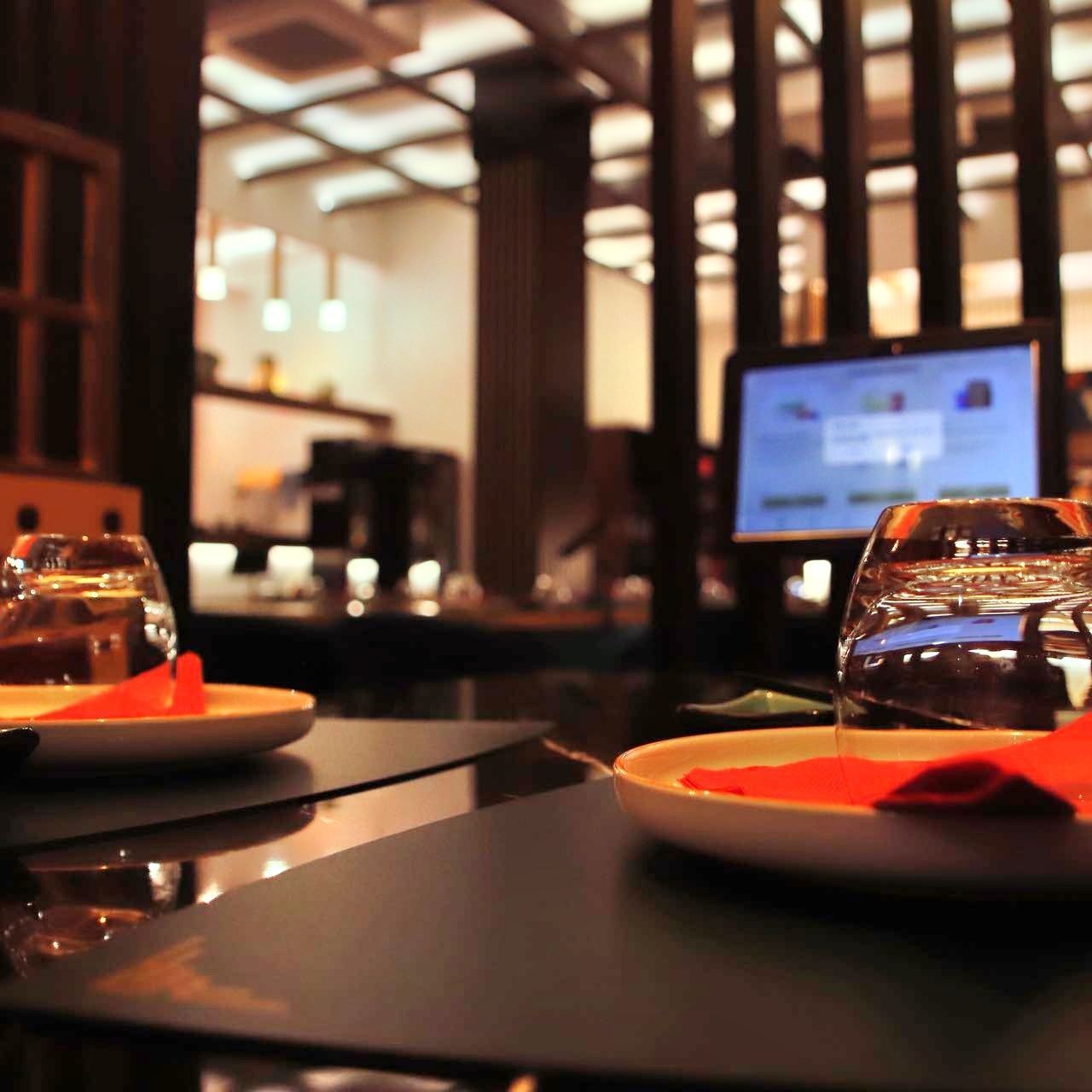 Tavolo del ristorante apparecchiato con calici da vino e tovaglioli arancioni, interno illuminato da una luce soffusa sullo sfondo.