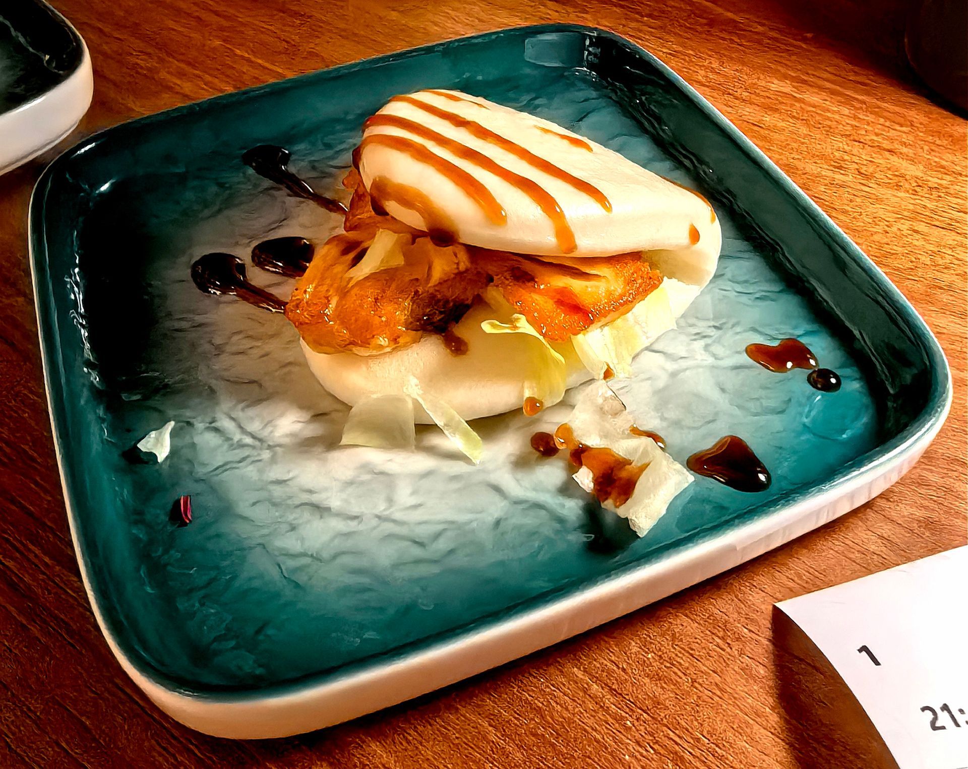 Dessert servito su un piatto color verde acqua con crema bianca e salsa al caramello su un tavolo di legno.