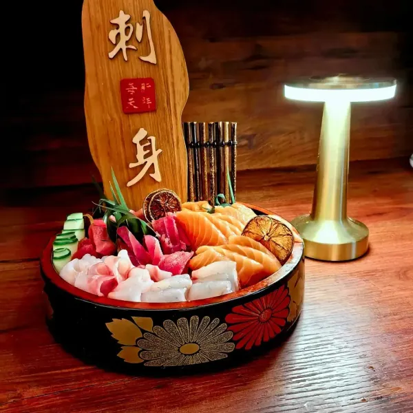 Piatto di sashimi in una ciotola decorativa su un tavolo di legno, illuminato da una piccola lampada, con una targhetta di legno alle sue spalle.