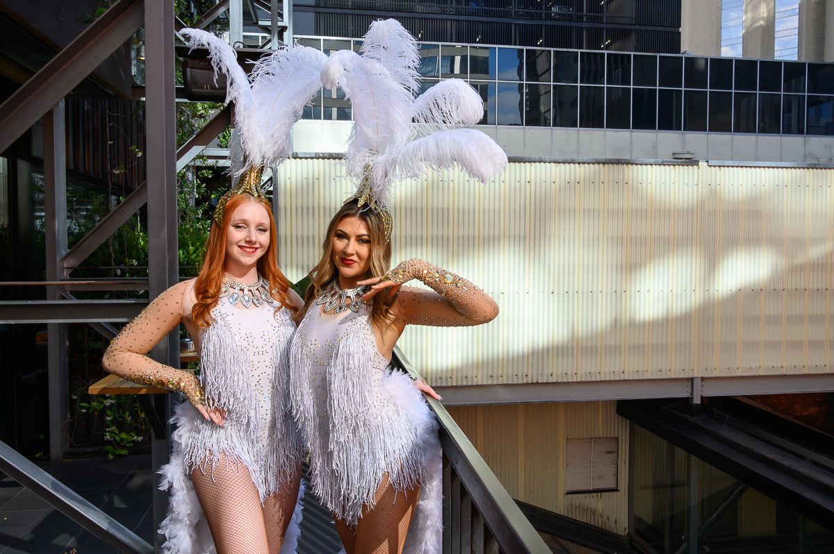 Two performers in white sequined fringe outfits and feathered headpieces pose together on a balcony.