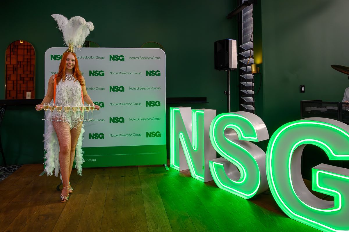A performer in a white feather costume stands by a backdrop and large, illuminated green 