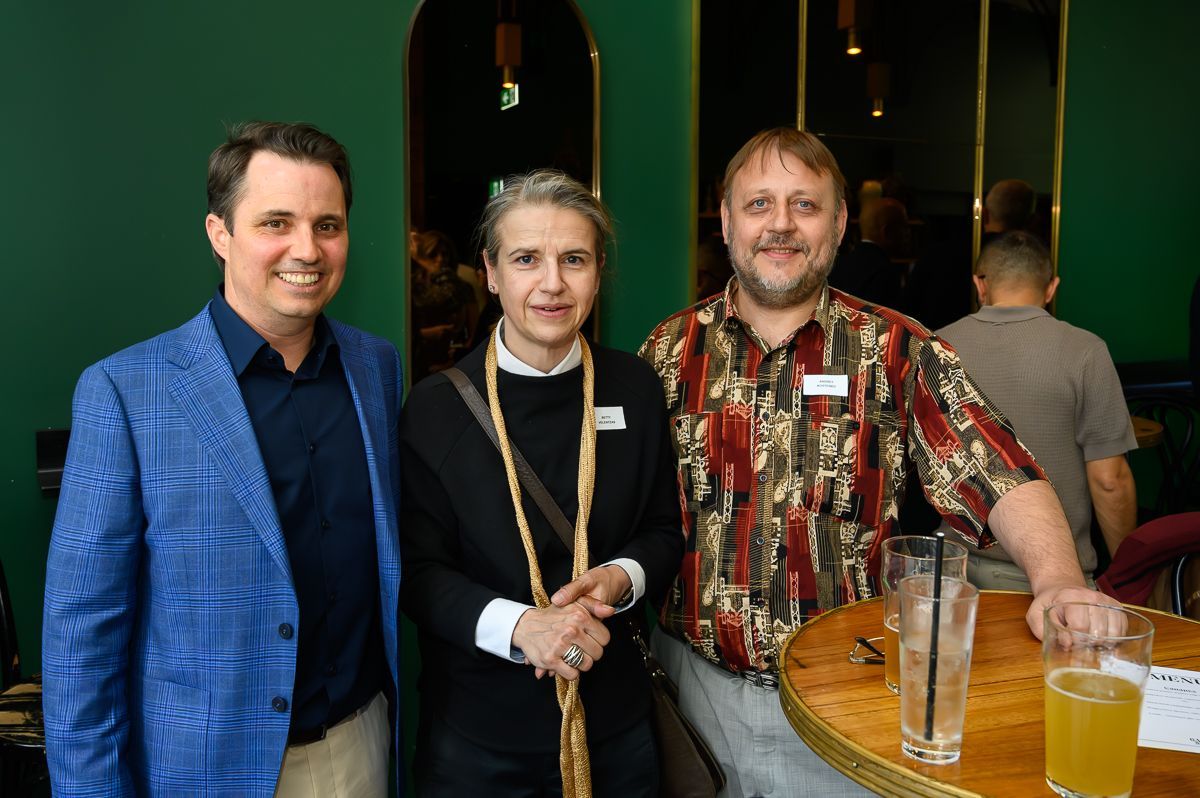 Three people pose together at a bar with green walls, two smiling toward the camera and one facing partially away.