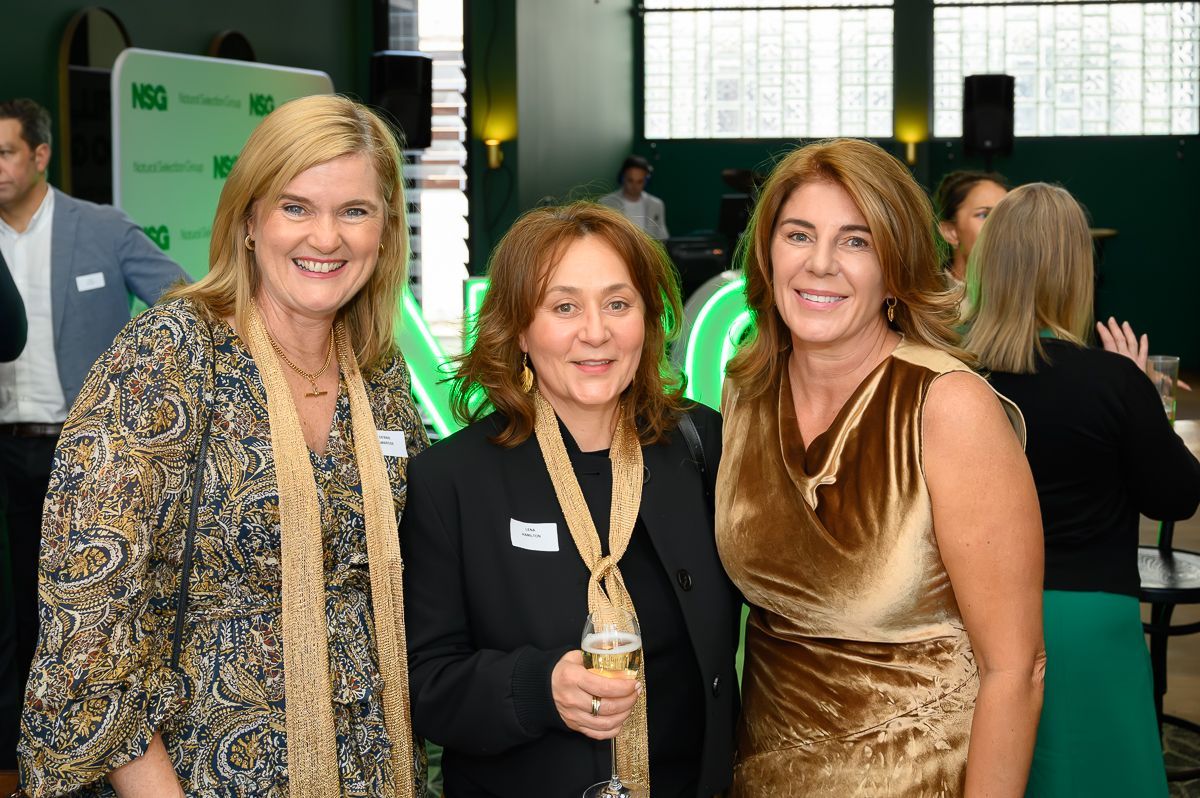 Three smiling people stand close together in an indoor event space, wearing semi-formal attire with scarves and dresses.
