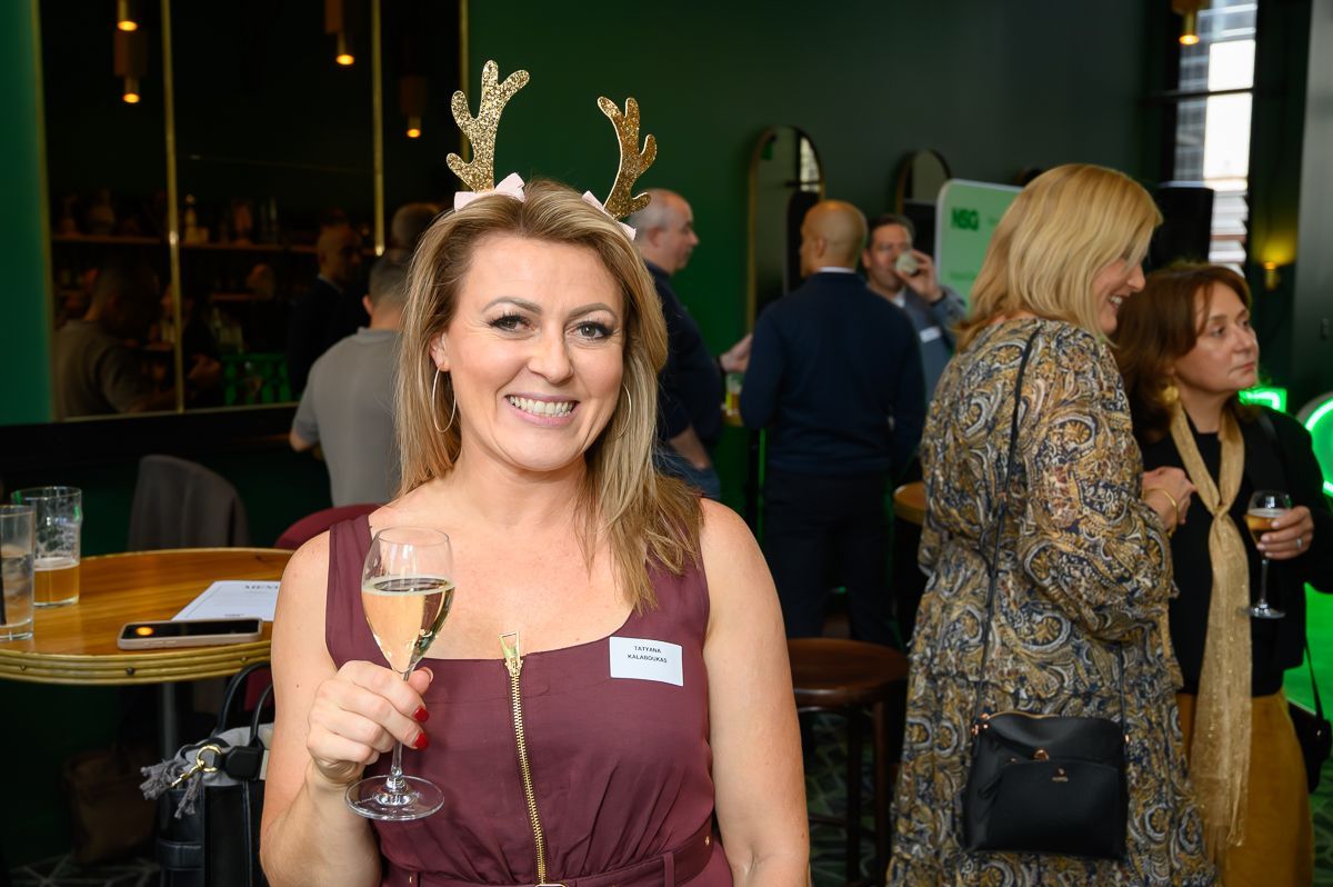 A smiling woman wearing gold reindeer antlers holds a glass of sparkling wine at an indoor social gathering.