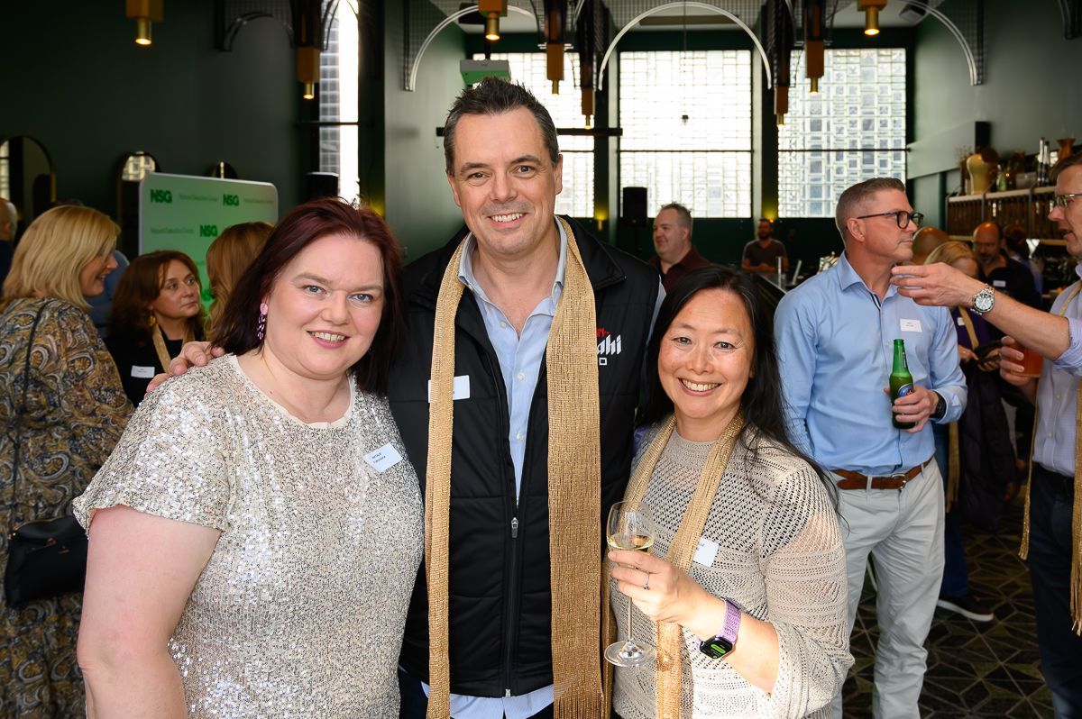 Three people stand side-by-side and smile at a social event, with blurred guests and a bright interior in the background.