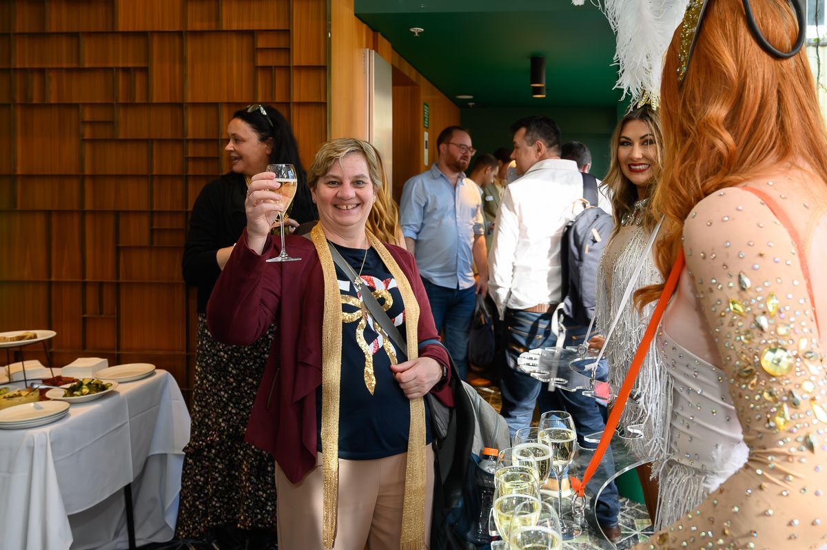 A smiling woman in a maroon cardigan and gold scarf raises a glass of champagne at an indoor gathering.