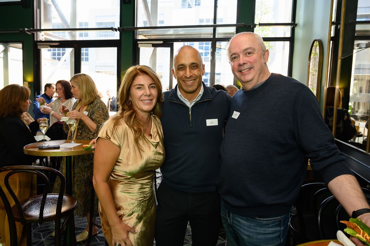 Three people stand smiling together at an indoor social gathering, with other attendees visible in the background.
