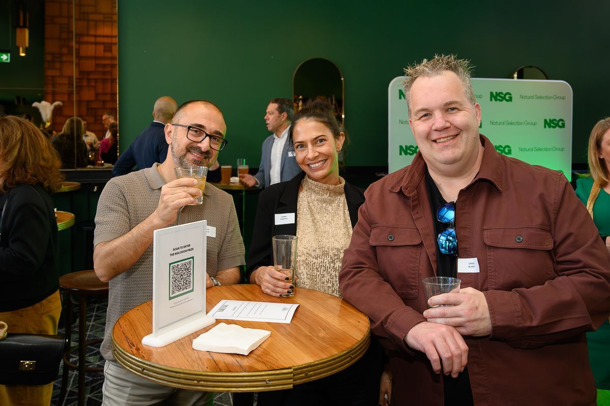 Three people smile while holding drinks at a round table with a QR code sign, in a room with green walls.