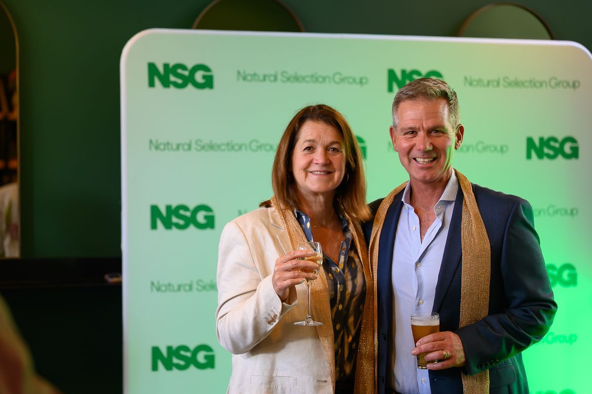 Two people smiling and holding drinks in front of a green Natural Selection Group backdrop.